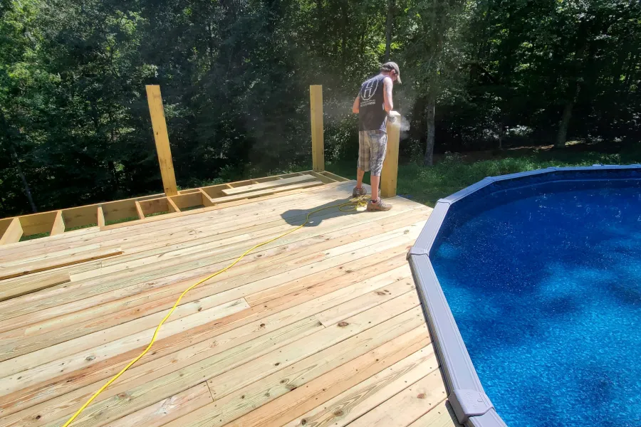 A man in a baseball cap and plaid shorts is sanding a wooden deck post beside a blue swimming pool.
